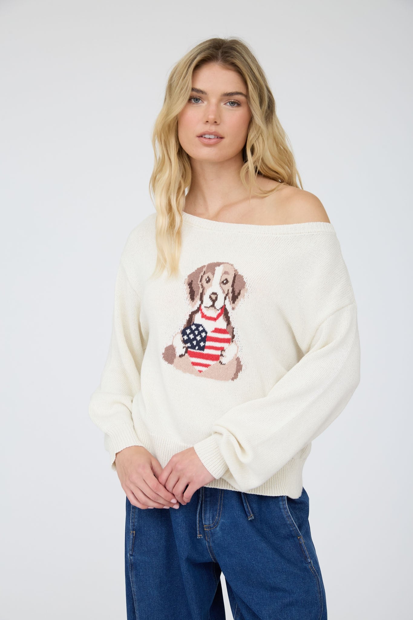 Cream knit sweater with an off-the-shoulder neckline and an American flag dog graphic on the front, styled with dark wash denim.