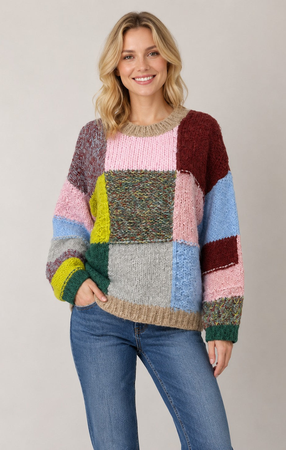 A woman wearing a chunky knit patchwork sweater with multiple colors including pink, blue, green, yellow, brown, and gray, paired with blue jeans, against a solid light gray background.