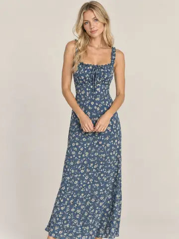 A woman modeling a sleeveless midi dress with a blue floral print, featuring thin straps, a gathered sweetheart neckline, and a flared skirt, set against a plain beige studio background.