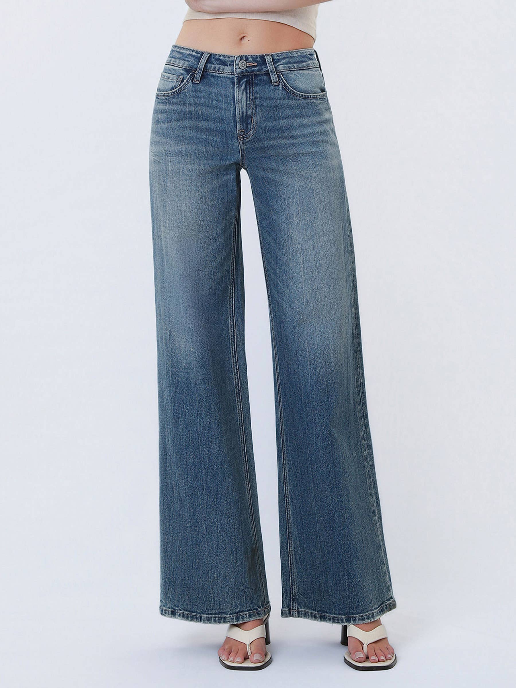 Model wearing high-rise wide-leg jeans in a vintage-inspired wash, styled with a cropped neutral tank and heeled sandals against a white studio background.