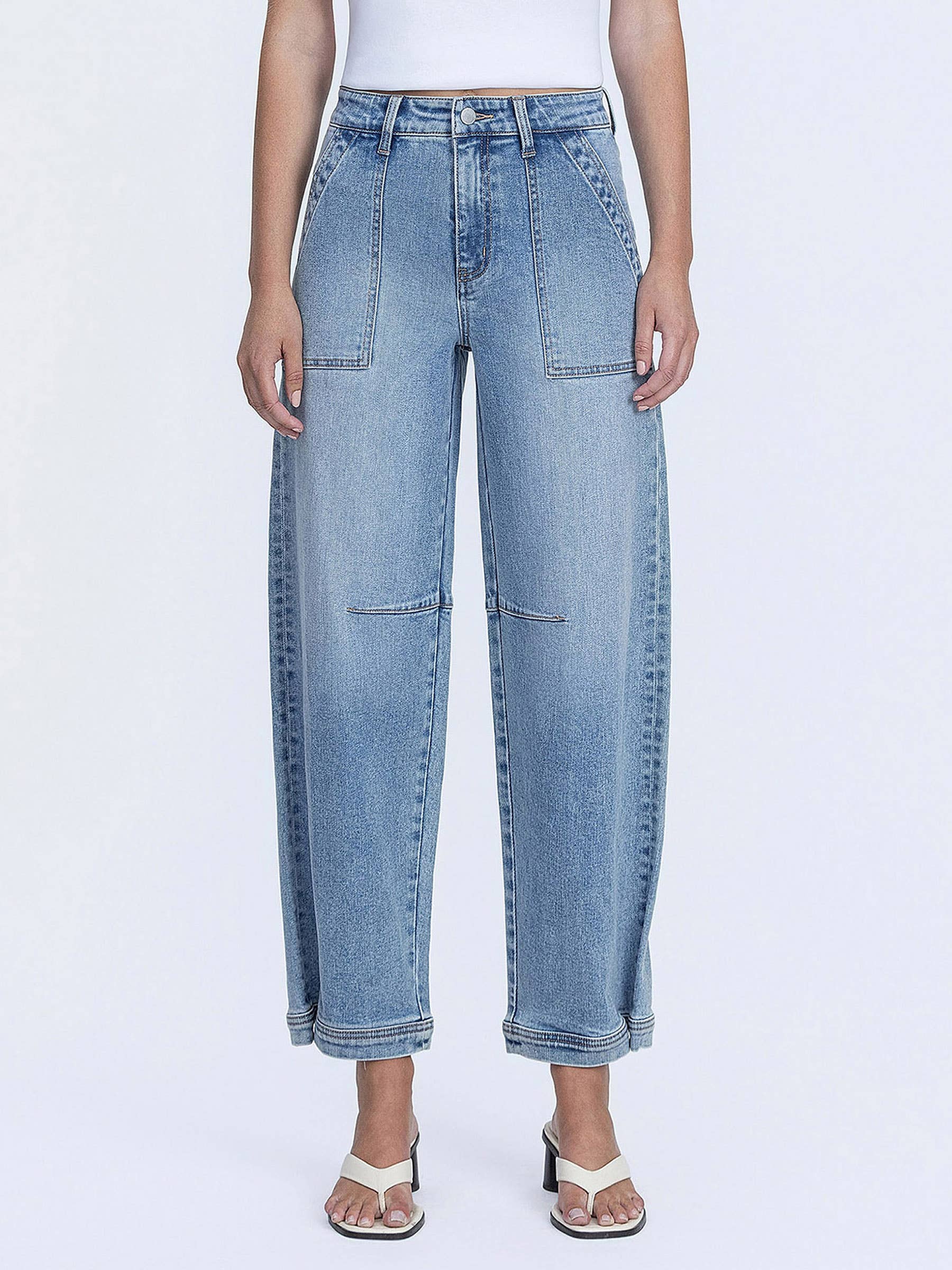 Model wearing high-rise barrel-leg jeans with oversized front patch pockets and a relaxed silhouette, styled with a fitted white bodysuit and heeled sandals against a white studio background.