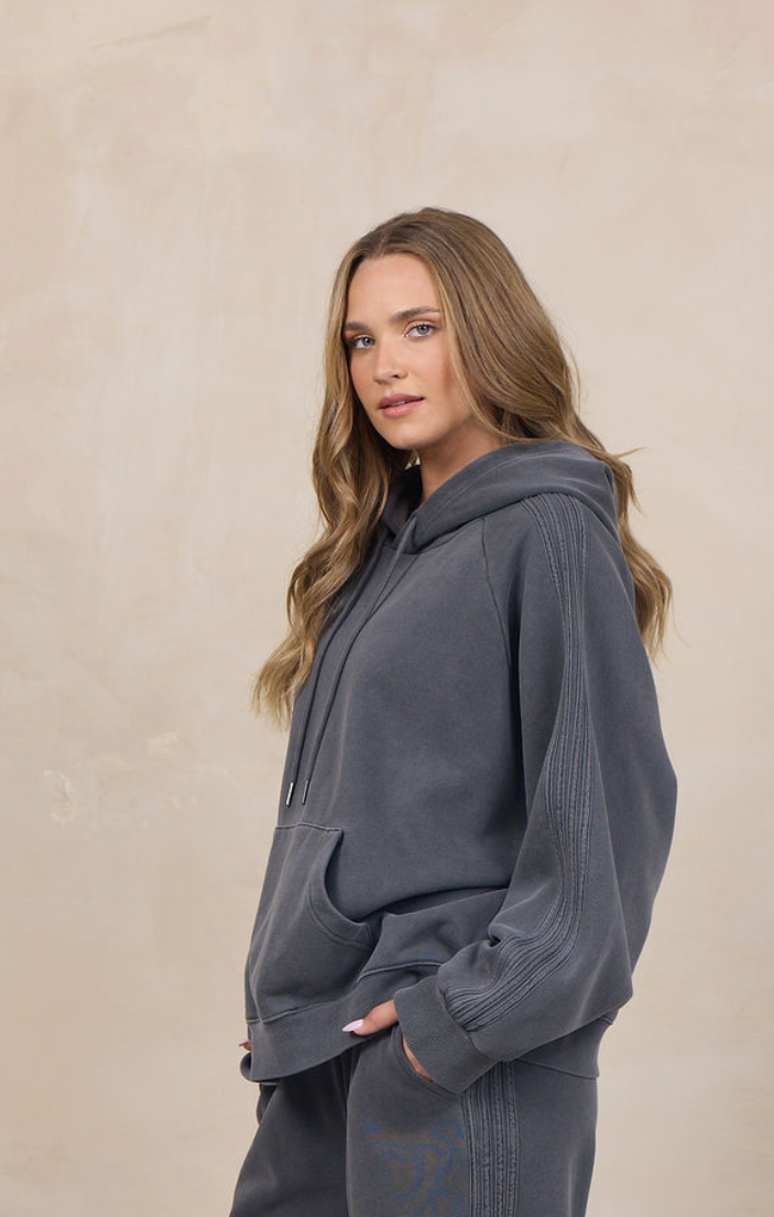 A woman modeling a dark gray two-piece loungewear set consisting of a pullover hoodie and matching wide-leg sweatpants, standing against a textured beige studio backdrop.