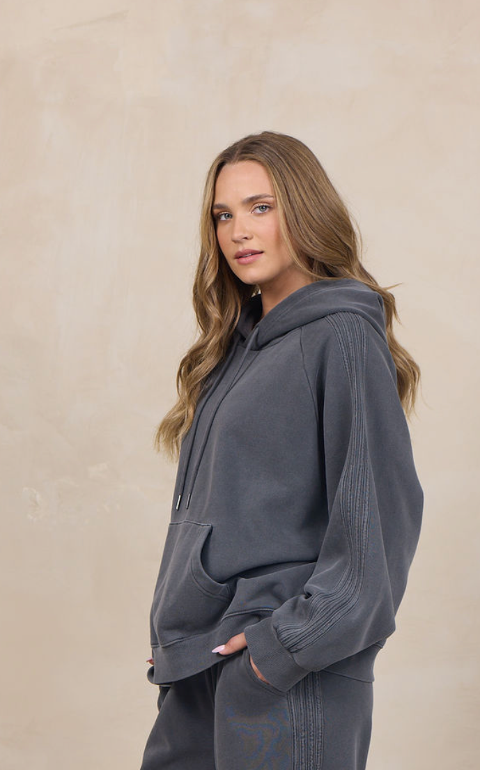 A woman modeling a dark gray two-piece loungewear set consisting of a pullover hoodie and matching wide-leg sweatpants, standing against a textured beige studio backdrop.