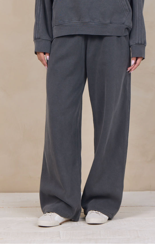 A woman modeling a dark gray matching lounge set consisting of a pullover hoodie and wide-leg sweatpants, standing against a textured beige studio backdrop.