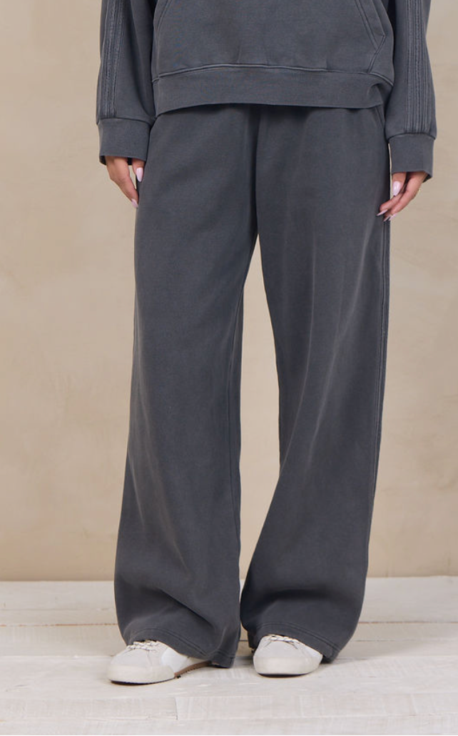A woman modeling a dark gray matching lounge set consisting of a pullover hoodie and wide-leg sweatpants, standing against a textured beige studio backdrop.