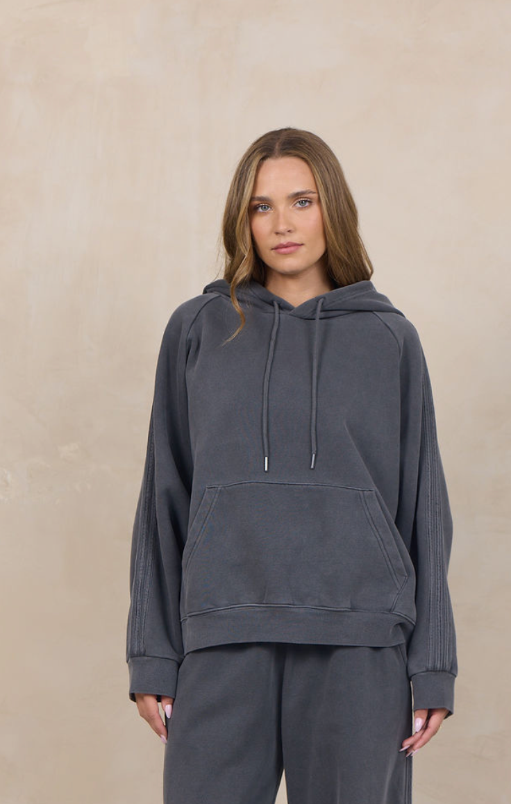 A woman modeling a dark gray matching lounge set consisting of a pullover hoodie and wide-leg sweatpants, standing against a textured beige studio backdrop.