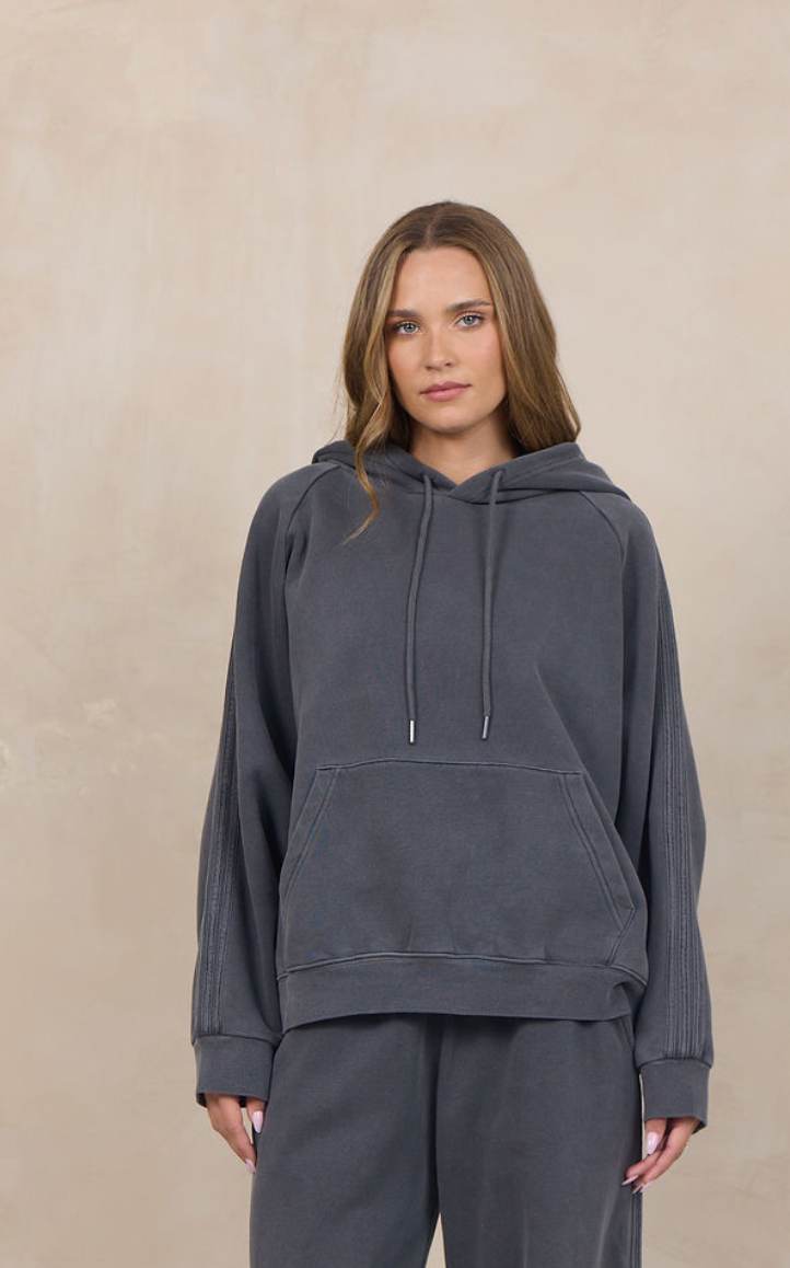 A woman modeling a dark gray matching lounge set consisting of a pullover hoodie and wide-leg sweatpants, standing against a textured beige studio backdrop.