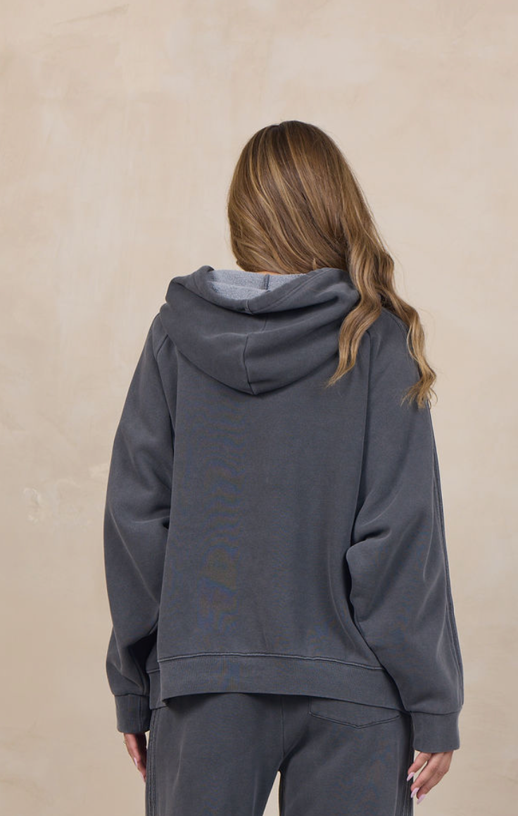 A woman viewed from behind wearing a dark gray matching lounge set consisting of a hooded sweatshirt and wide-leg sweatpants, standing against a textured beige wall.