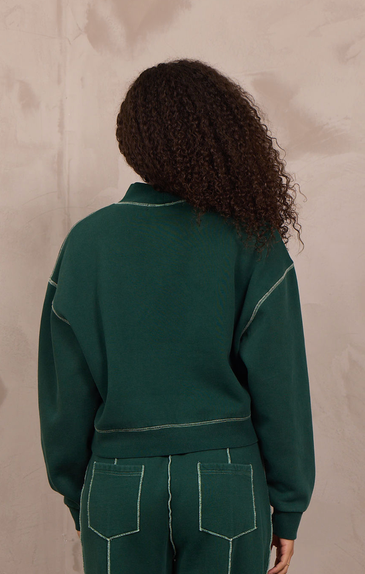 A model viewed from behind wearing a dark green two-piece tracksuit with contrasting light green stitching, consisting of a cropped long-sleeve top and matching wide-leg pants, standing against a textured beige wall.