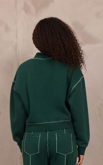 A model viewed from behind wearing a dark green two-piece tracksuit with contrasting light green stitching, consisting of a cropped long-sleeve top and matching wide-leg pants, standing against a textured beige wall.