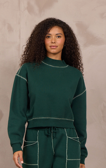 A woman modeling a matching dark green sweatshirt and wide-leg sweatpants set with contrasting light stitching, standing against a textured beige wall.
