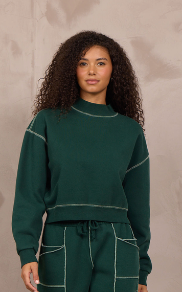 A woman modeling a matching dark green sweatshirt and wide-leg sweatpants set with contrasting light stitching, standing against a textured beige wall.
