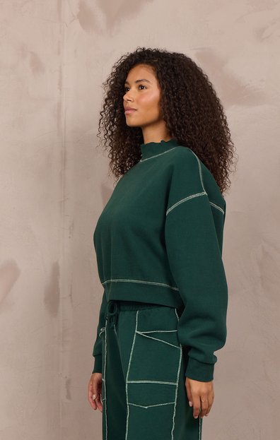 A woman stands in profile wearing a dark green knit matching set consisting of a long-sleeve cropped sweatshirt and wide-leg pants with contrasting light beige stitching. She is wearing beige sneakers and standing against a textured, warm-toned concrete wall.