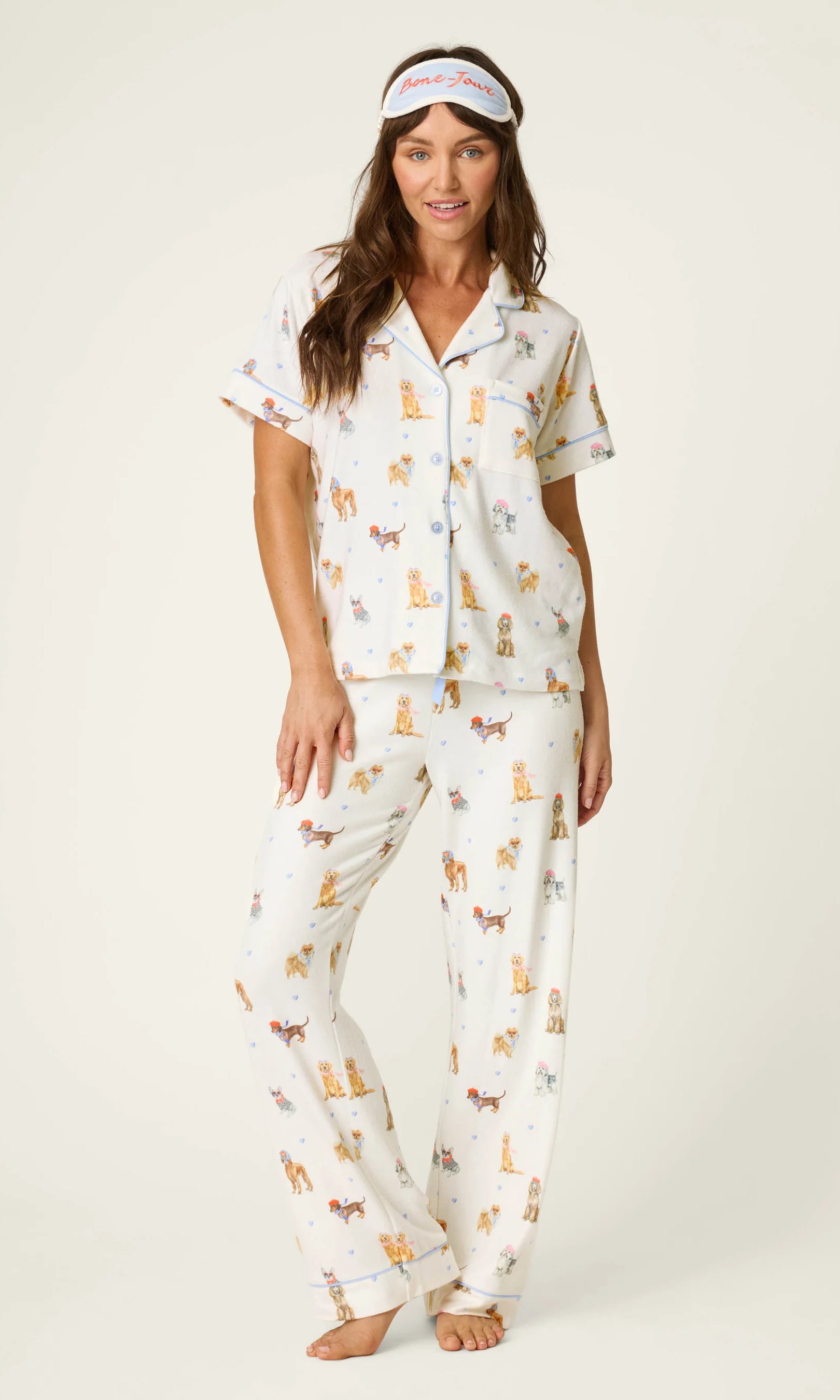 Woman wearing a three-piece dog-print pajama set with a button-front top, relaxed pants, and matching sleep mask.