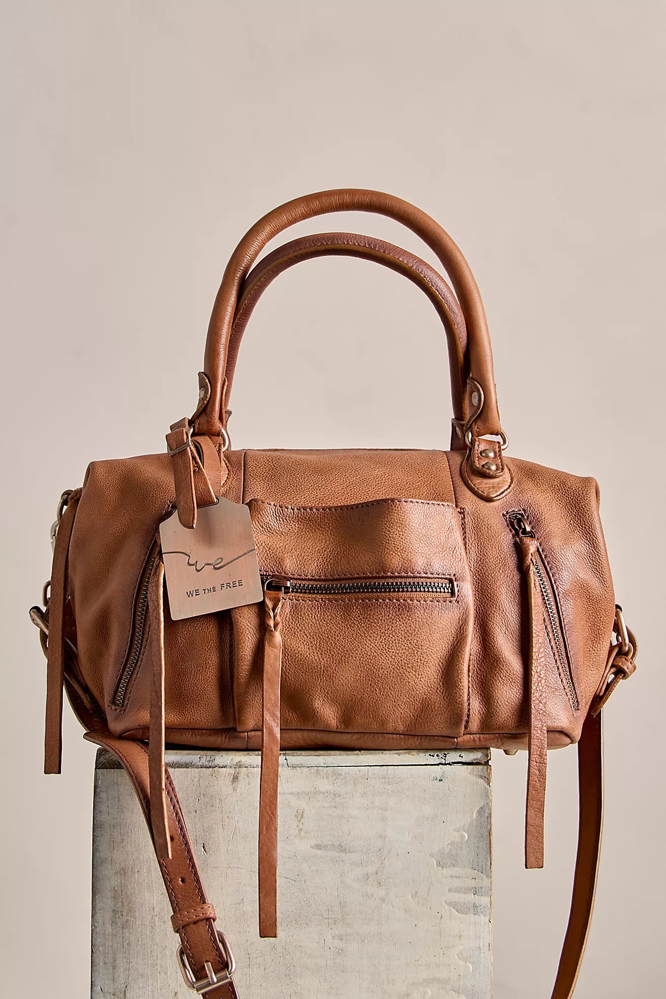 Leather satchel handbag with dual top handles, multiple exterior zip pockets, and a removable crossbody strap displayed on a pedestal against a neutral background.