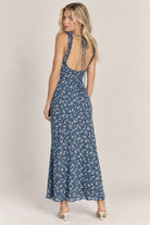A woman modeling a sleeveless, open-back maxi dress with a blue and white floral pattern, photographed against a plain light beige studio background.