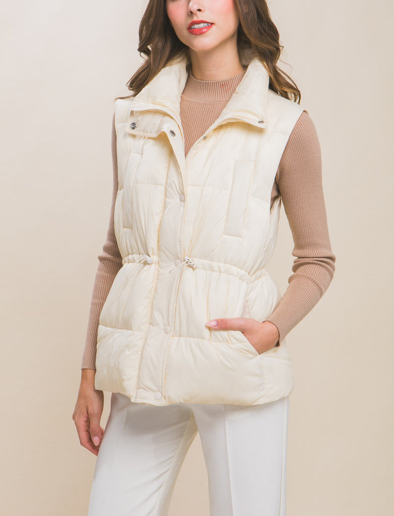 Moonbeam Zip Up Button Puffer Vest Cream. This zip up puffer vest features waist toggles and side pockets, the perfect layering piece to keep you warm in the chillier weather all season long.