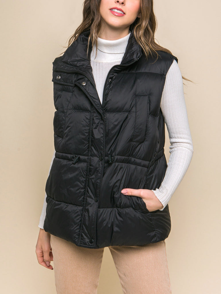 Moonbeam Zip Up Button Puffer Vest Black. This zip up puffer vest features waist toggles and side pockets, the perfect layering piece to keep you warm in the chillier weather all season long.