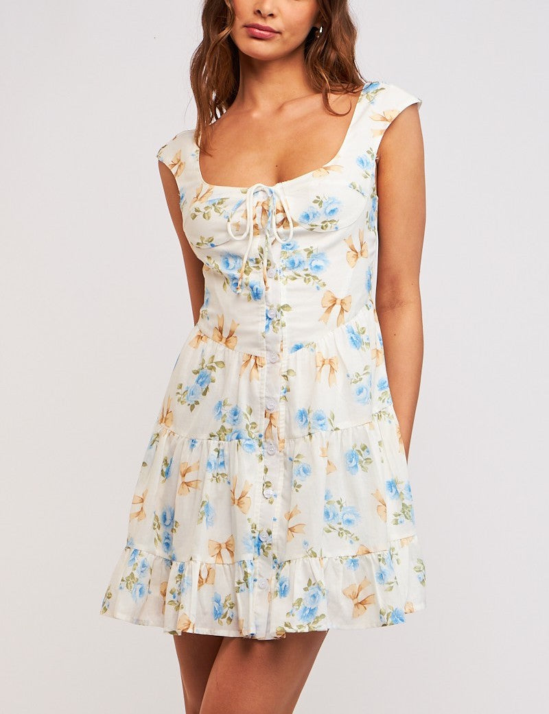 Tessa Bow Print Tie Front Dress Blue Ribbon Floral. This printed mini dress features a u-neck with wide straps and a button front design with a tie detail, the cutest piece to add to your spring wardrobe.