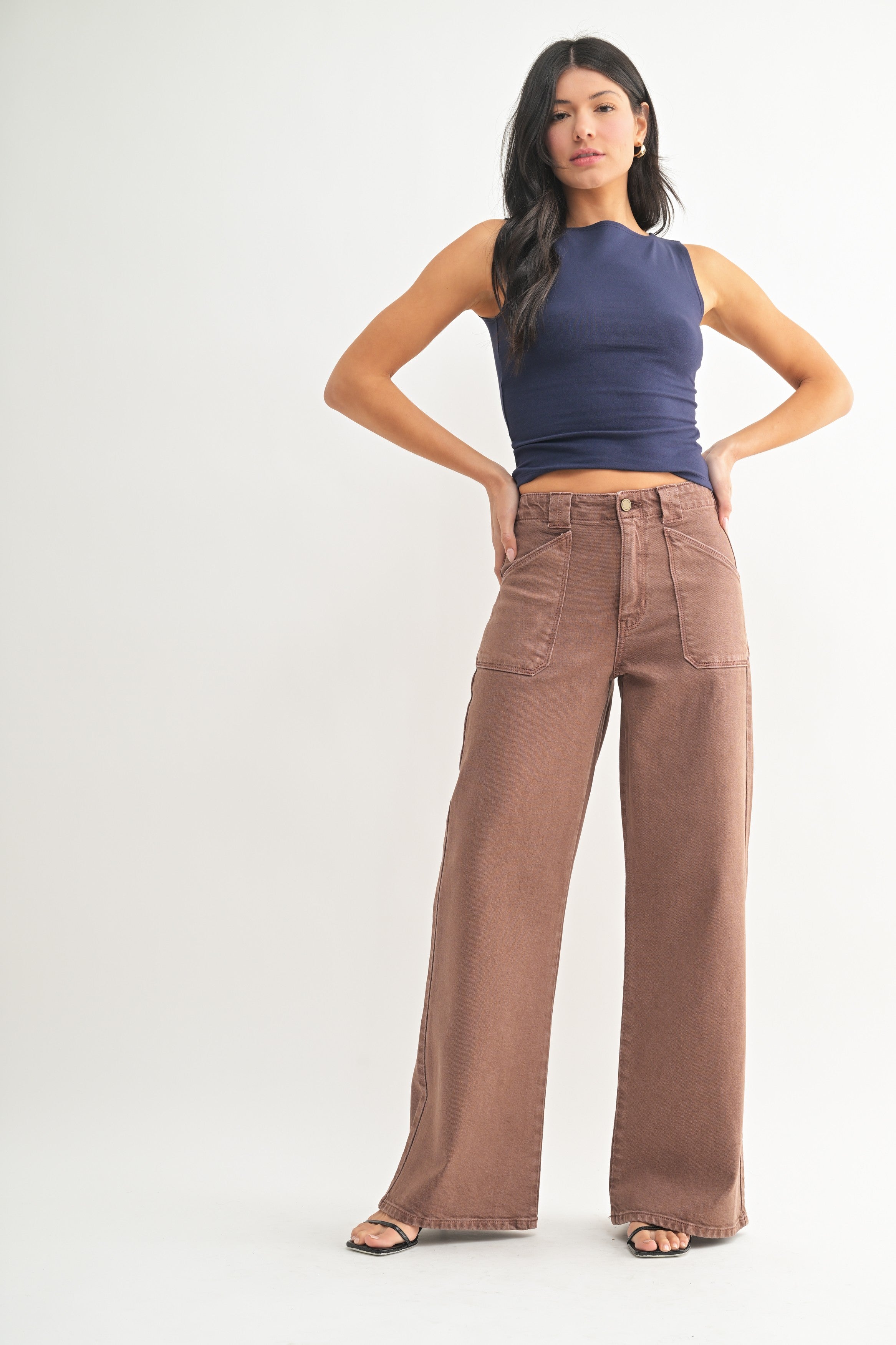 A woman modeling a pair of high-waisted, wide-leg brown pants with a navy blue sleeveless crop top, standing against a plain light gray studio background.
