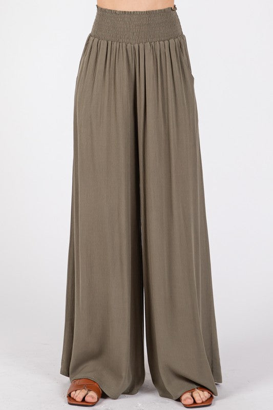 Model wearing flowy wide-leg pants with a smocked waistband, side pockets, and a relaxed fit.