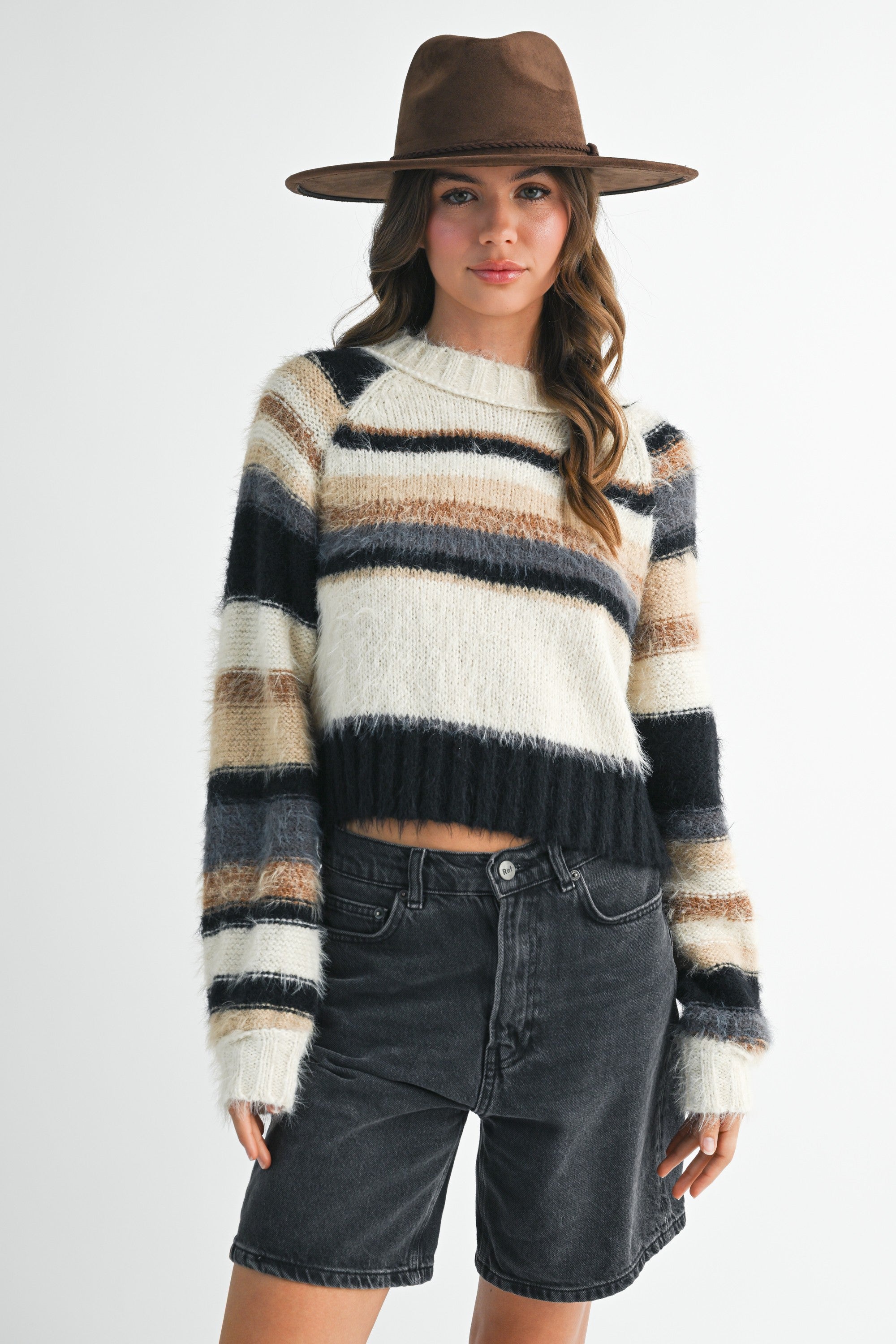 A woman wearing a fuzzy striped knit sweater with horizontal bands of cream, beige, brown, and black, paired with dark denim shorts and a brown wide-brim hat, against a light gray studio background.
