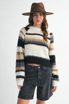 A woman wearing a fuzzy striped knit sweater with horizontal bands of cream, beige, brown, and black, paired with dark denim shorts and a brown wide-brim hat, against a light gray studio background.