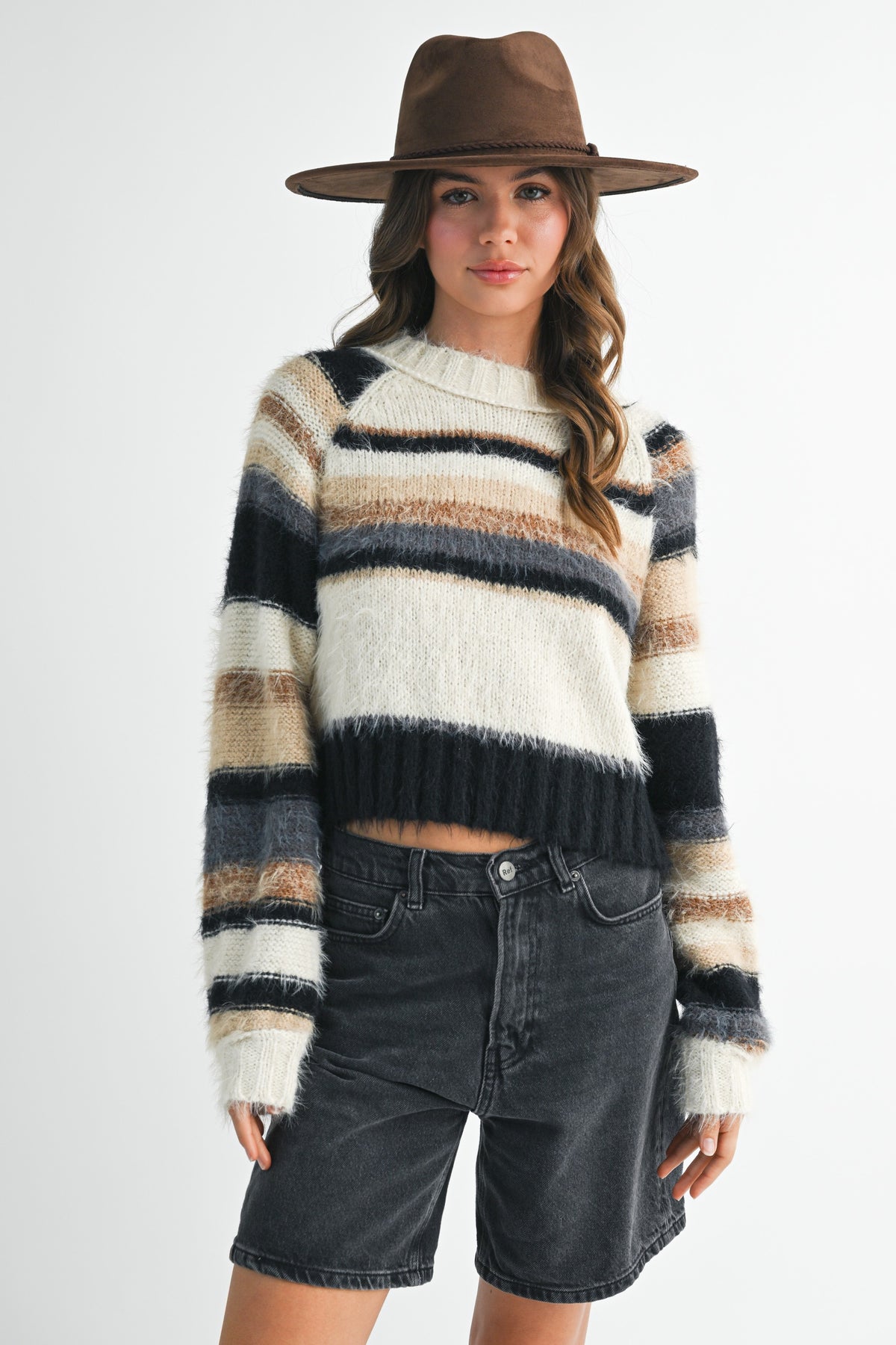 A woman wearing a fuzzy striped knit sweater with horizontal bands of cream, beige, brown, and black, paired with dark denim shorts and a brown wide-brim hat, against a light gray studio background.