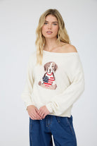 Cream knit sweater with an off-the-shoulder neckline and an American flag dog graphic on the front, styled with dark wash denim.