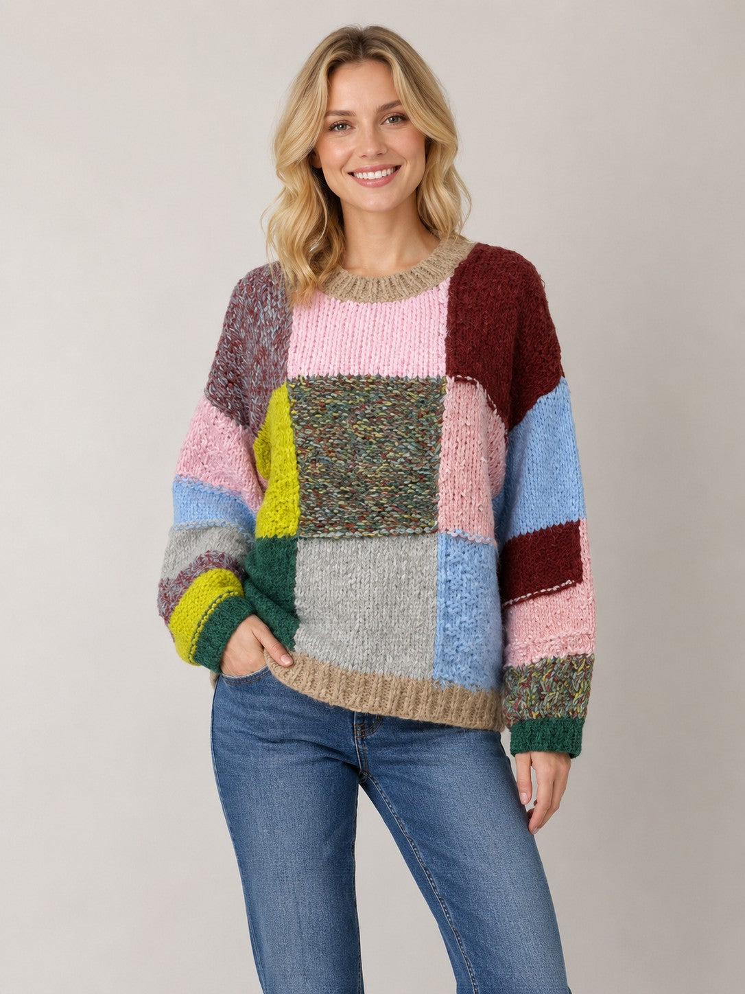 A woman wearing a chunky knit patchwork sweater with multiple colors including pink, blue, green, yellow, brown, and gray, paired with blue jeans, against a solid light gray background.