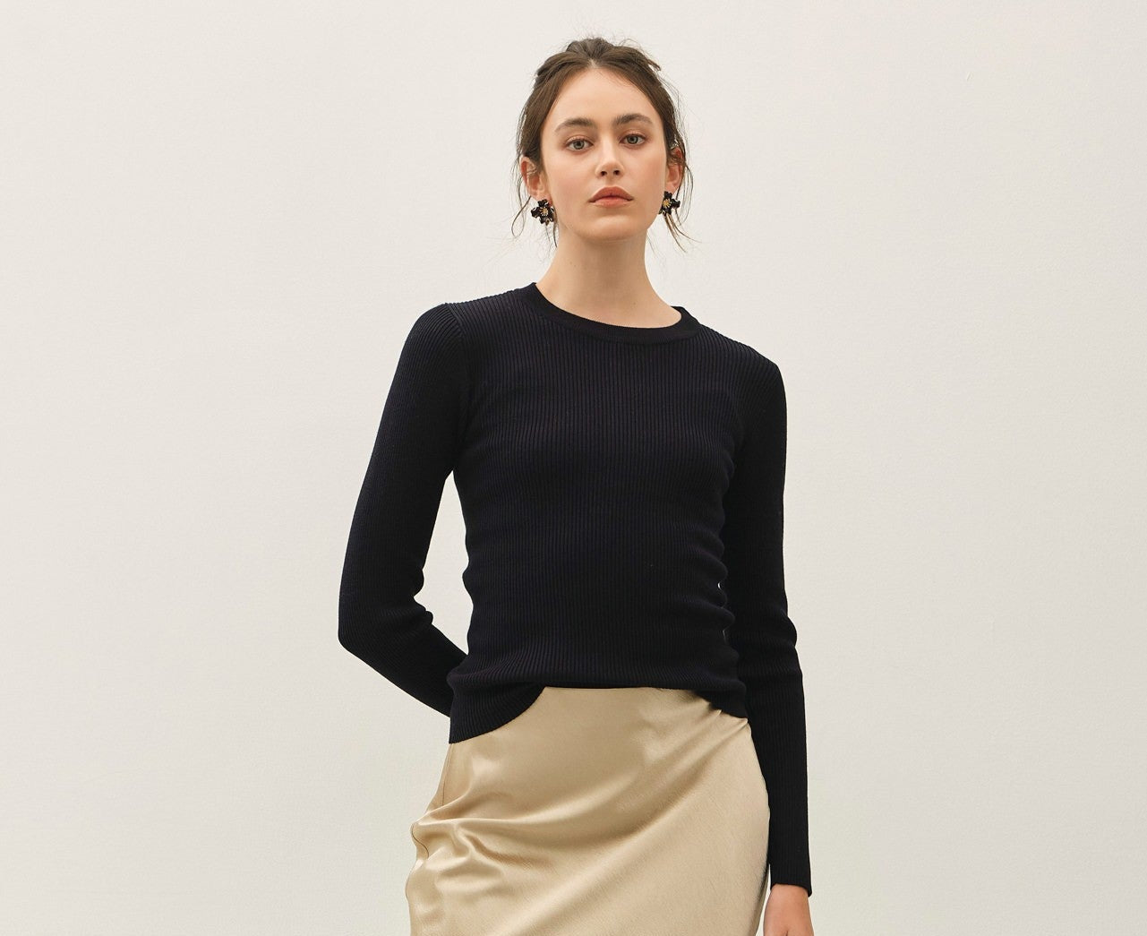 A woman modeling a black long-sleeve knit top and a beige satin midi skirt with side slits, paired with black strappy heeled sandals, against a plain off-white studio background.