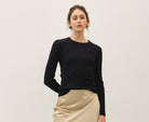 A woman modeling a black long-sleeve knit top and a beige satin midi skirt with side slits, paired with black strappy heeled sandals, against a plain off-white studio background.