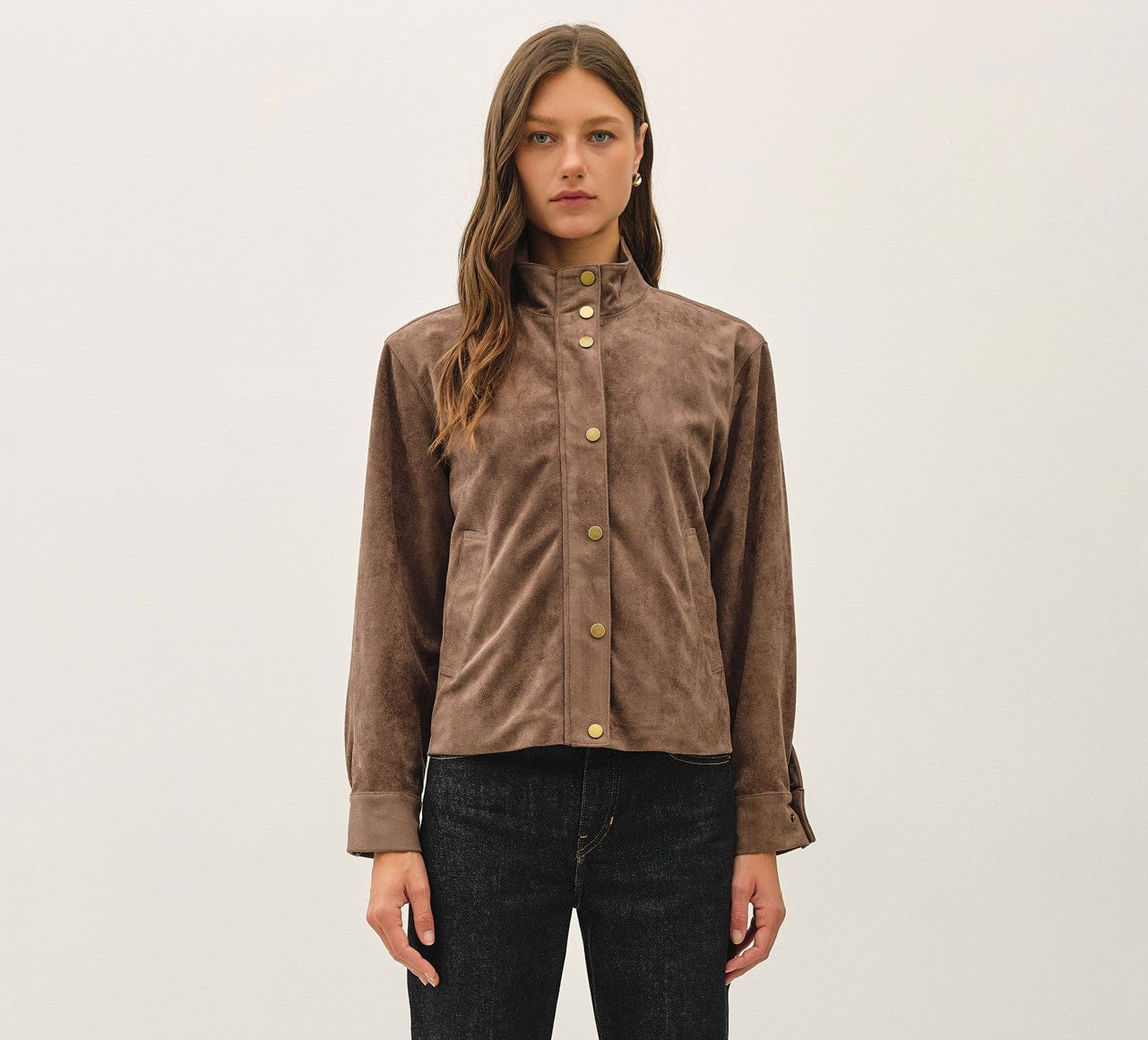 A woman stands against a plain white studio background wearing a brown corduroy jacket with gold-tone buttons and dark charcoal gray jeans.