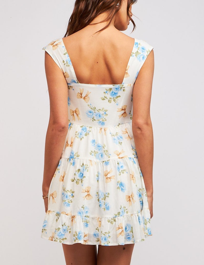 Tessa Bow Print Tie Front Dress Blue Ribbon Floral. This printed mini dress features a u-neck with wide straps and a button front design with a tie detail, the cutest piece to add to your spring wardrobe.