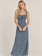 A woman modeling a sleeveless midi dress with a blue floral print, featuring thin straps, a gathered sweetheart neckline, and a flared skirt, set against a plain beige studio background.