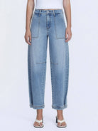 Model wearing high-rise barrel-leg jeans with oversized front patch pockets and a relaxed silhouette, styled with a fitted white bodysuit and heeled sandals against a white studio background.