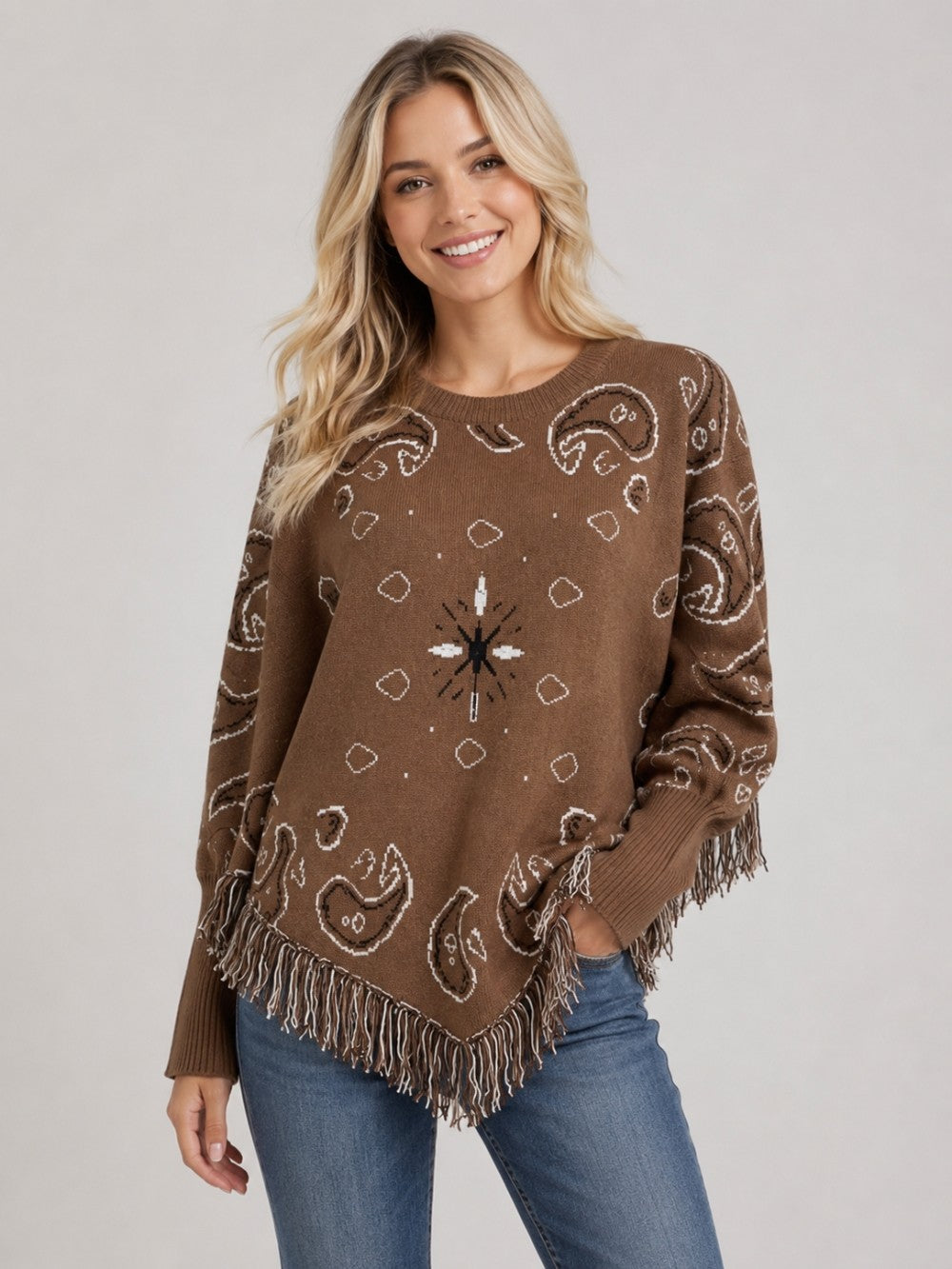 A woman wearing a brown knit poncho sweater with white paisley and geometric patterns, fringed hem and sleeves, paired with blue jeans, against a light gray studio background.