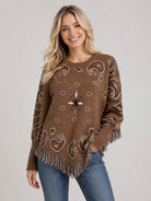 A woman wearing a brown knit poncho sweater with white paisley and geometric patterns, fringed hem and sleeves, paired with blue jeans, against a light gray studio background.