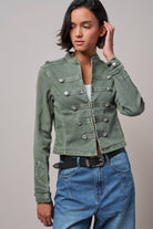 A woman modeling a cropped, double-breasted military-style jacket in sage green. The jacket features silver-toned snap buttons and flap pockets, worn over a white top and blue jeans against a solid gray studio background.