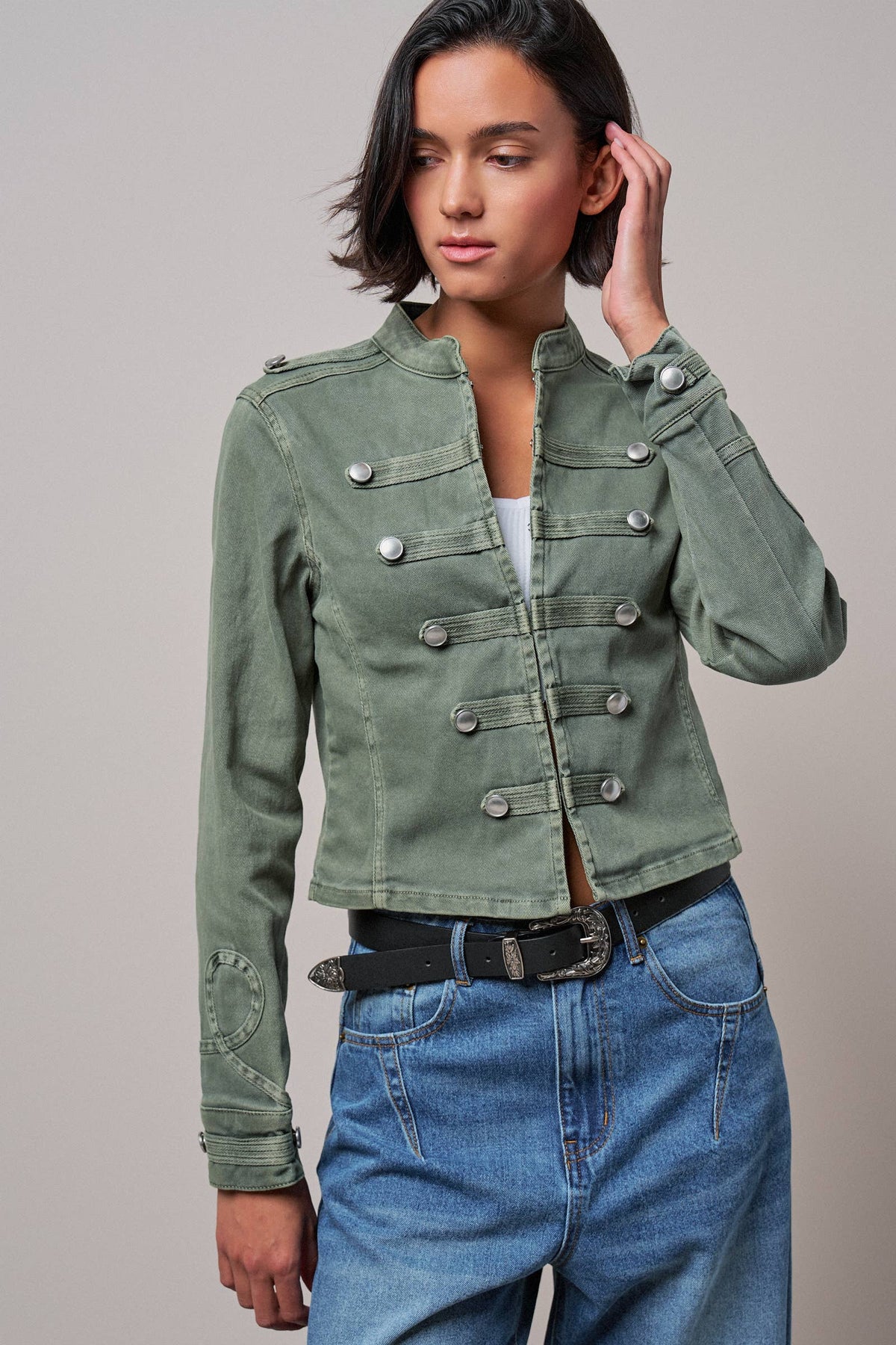 A woman modeling a cropped, double-breasted military-style jacket in sage green. The jacket features silver-toned snap buttons and flap pockets, worn over a white top and blue jeans against a solid gray studio background.