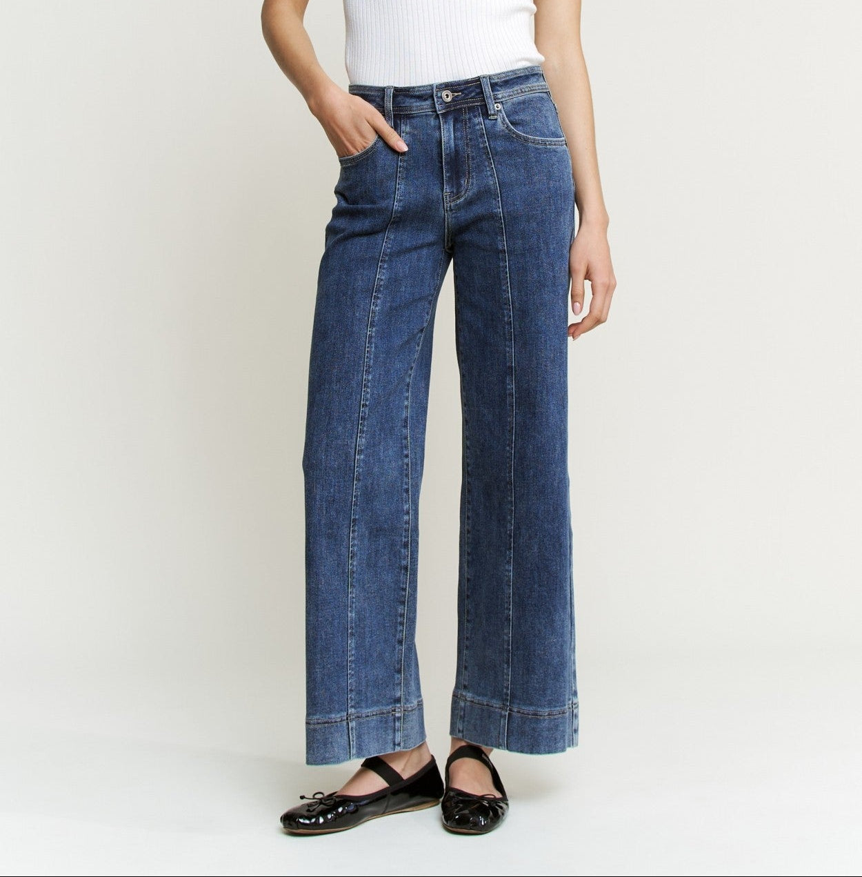 A woman modeling a white short-sleeve crewneck top and high-waisted, wide-leg blue denim jeans with black flat shoes, standing against a plain light gray studio background.