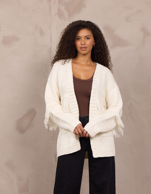 Woman wearing an oversized open-front cable knit cardigan with fringe trim and ribbed cuffs.