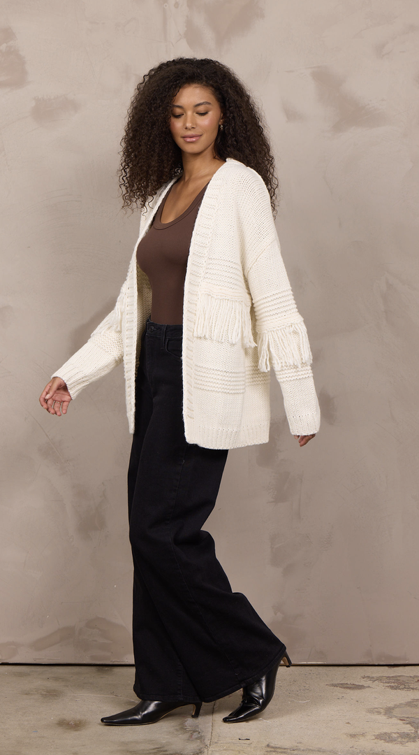 Woman wearing an oversized open-front cable knit cardigan with fringe trim and ribbed cuffs.