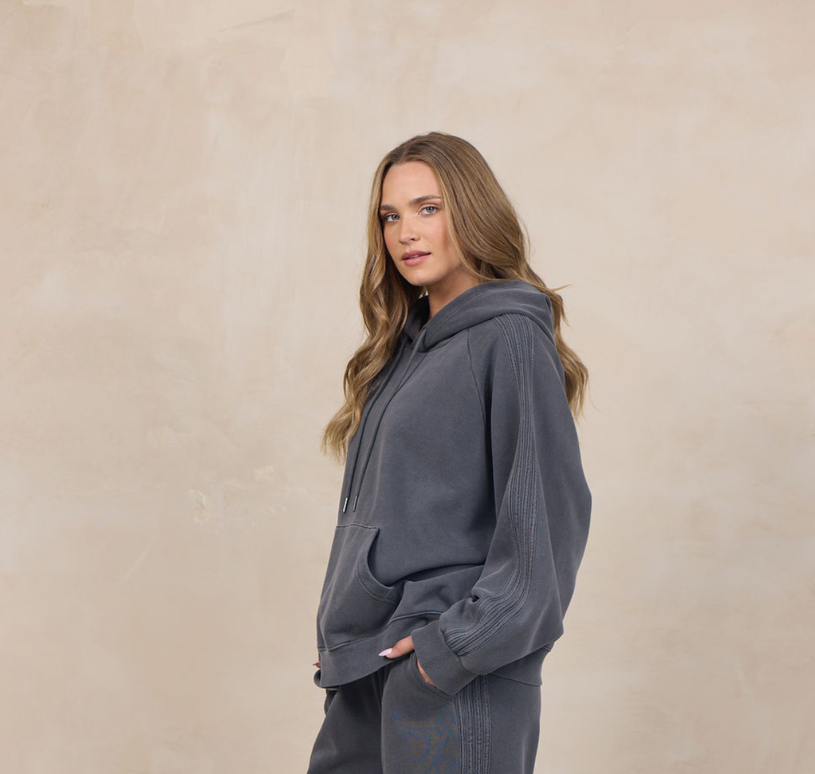 A woman modeling a dark gray two-piece loungewear set consisting of a pullover hoodie and matching wide-leg sweatpants, standing against a textured beige studio backdrop.