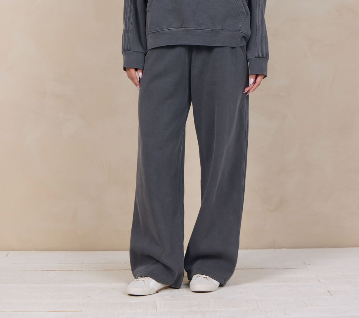 A woman modeling a dark gray matching lounge set consisting of a pullover hoodie and wide-leg sweatpants, standing against a textured beige studio backdrop.