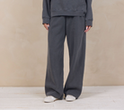 A woman modeling a dark gray matching lounge set consisting of a pullover hoodie and wide-leg sweatpants, standing against a textured beige studio backdrop.