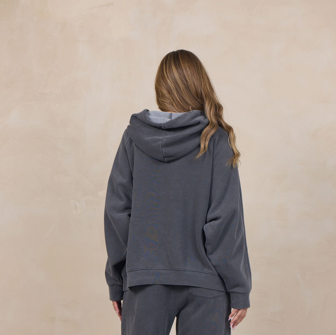 A woman viewed from behind wearing a dark gray matching lounge set consisting of a hooded sweatshirt and wide-leg sweatpants, standing against a textured beige wall.