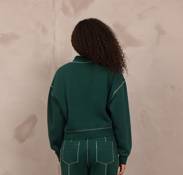 A model viewed from behind wearing a dark green two-piece tracksuit with contrasting light green stitching, consisting of a cropped long-sleeve top and matching wide-leg pants, standing against a textured beige wall.