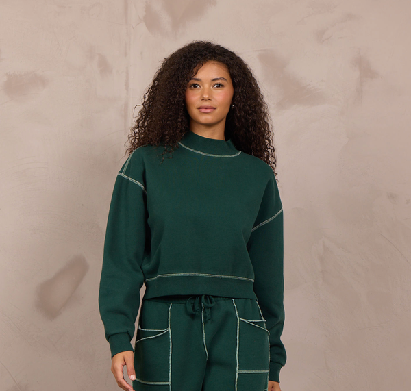 A woman modeling a matching dark green sweatshirt and wide-leg sweatpants set with contrasting light stitching, standing against a textured beige wall.