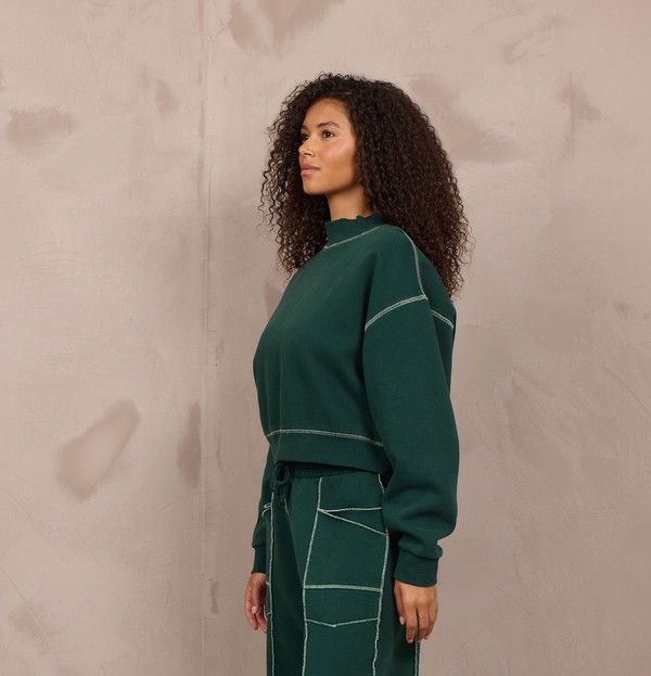 A woman stands in profile wearing a dark green knit matching set consisting of a long-sleeve cropped sweatshirt and wide-leg pants with contrasting light beige stitching. She is wearing beige sneakers and standing against a textured, warm-toned concrete wall.